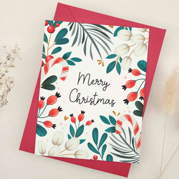 Christmas card on slightly textured off-white cardstock with “Merry Christmas” in black script, surrounded by greenery, red berries, cream leaves, and candy-cane accents, paired with envelope — XOXOKristen