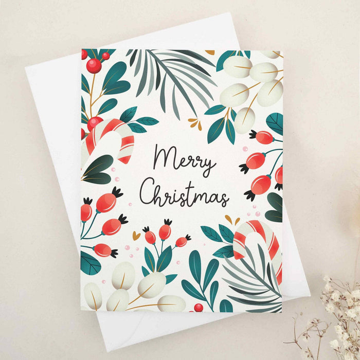 Folded holiday card with festive botanical border of pine leaves, holly berries, and cream foliage framing the words “Merry Christmas” — XOXOKristen.