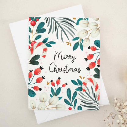 Folded holiday card with festive botanical border of pine leaves, holly berries, and cream foliage framing the words “Merry Christmas” — XOXOKristen.