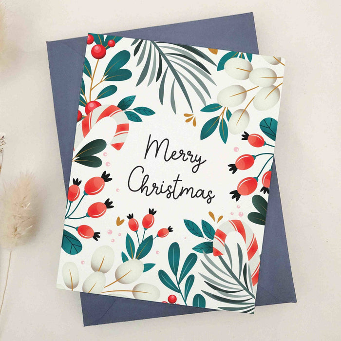 Standing card on textured off-white cardstock with botanical wreath-style border of leaves and berries framing “Merry Christmas,” displayed with coordinating envelope — XOXOKristen.